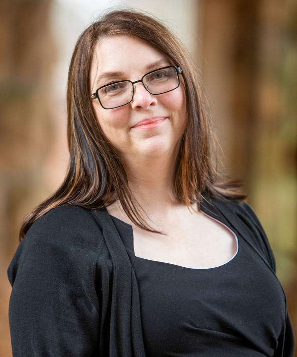 A white woman smiling with long brown hair and glasses, wearing a black blouse and cardigan.